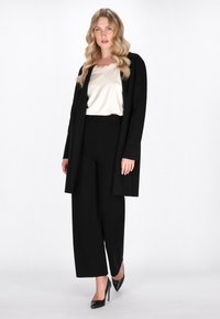 Zwarte lange cardigan over een crèmekleurige satijnen blouse, gecombineerd met zwarte wijde broek en zwarte hoge hakken. Soepele stof met een relaxte pasvorm.