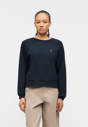 Jonge vrouw met gevlochten haar, gekleed in een donkerblauwe trui met een klein rood hartje op de borst en beige broek.