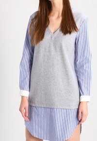 Top de mezcla de lana gris con escote en V, superpuesto sobre una camisa de rayas azul claro y blanca. Manga larga y detalle de dobladillo curvado.