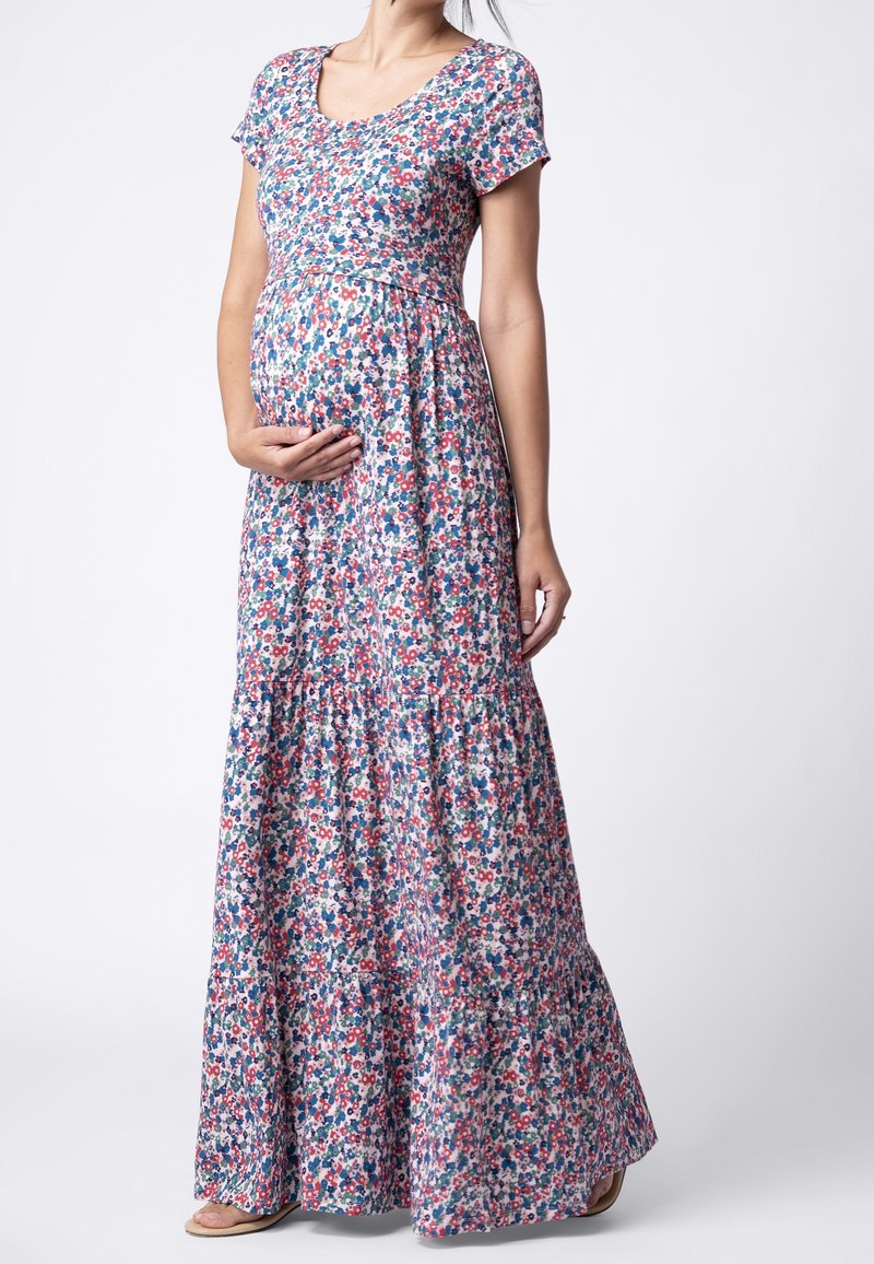 Femme enceinte portant une robe maxi à manches courtes à motif floral, tenant doucement son ventre, debout devant un fond clair uni.