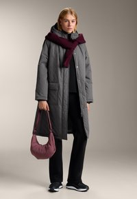 Šedý prešitý dlhý kabát s kapucňou, bordový pletený šál, čierny top, čierne nohavice a šedá taška, doplnené čiernymi teniskami.