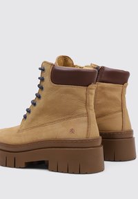 Botas de tobillo de cuero marrón claro con cuellos acolchados marrón oscuro, cordones negros, suelas robustas marrón con tacos y cremalleras laterales, mostradas desde ángulos trasero y lateral.