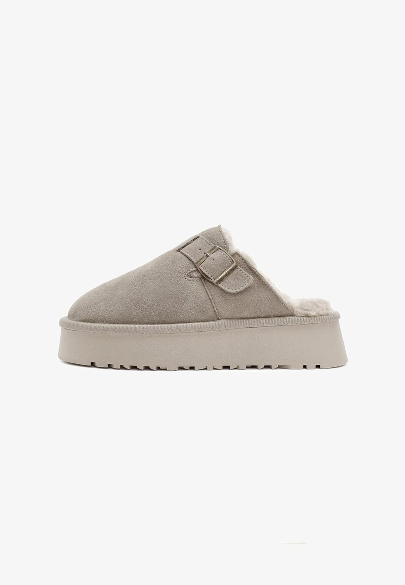 Zapato beige de ante de tipo slip-on con suela de goma gruesa, forro de piel texturizada y dos hebillas ajustables en la parte superior.