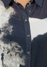 Nahaufnahme eines geknöpften navyblauen Hemdes mit abstraktem beigefarbenem und weißen Muster und spitzem Kragen.
