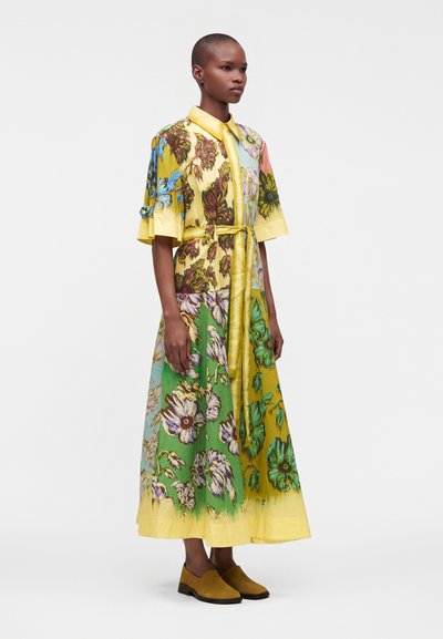 Femme portant une robe longue fleurie colorée avec col et manches jaunes, nouée à la taille, associée à des chaussures couleur moutarde.