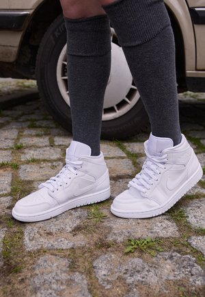 Białe wysokie sneakersy Nike noszone z ciemnoszarymi skarpetami do kolan, stojące na powierzchni z kostki brukowej obok beżowego koła samochodu.