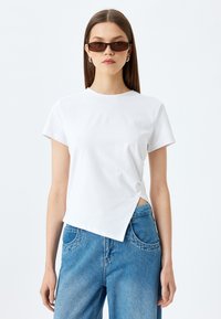 Camiseta blanca de manga corta con dobladillo curvado y una abertura lateral, combinada con jeans de mezclilla azul claro que tienen bolsillos redondeados.