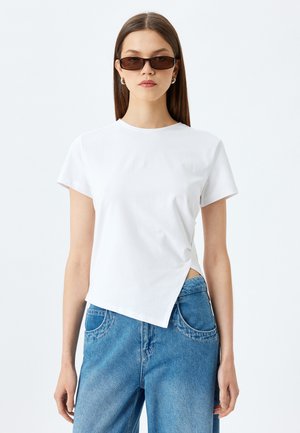 T-shirt blanc à manches courtes avec un ourlet arrondi et une fente sur le côté, associé à un jean en denim bleu clair avec des poches arrondies.