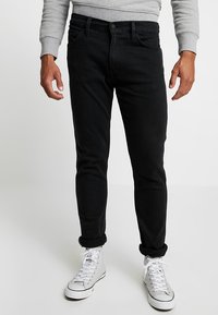 Jeans in denim nero slim fit, con un classico design a cinque tasche, orli arrotolati, indossati con sneakers bianche alte.