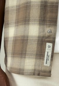 Manžeta béžové a hnědé kárované košile s kovovým drukem a bílou textilní štítkem s nápisem „Durable Goods Lot. 082“ na pozadí světlých kalhot.