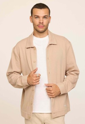 Veste beige boutonnée avec une texture douce, dotée d'un col et de poches avant, portée sur une chemise blanche. Tissu lisse et design décontracté.