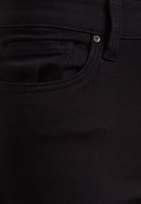 Jeans de mezclilla negra con una textura suave, corte ajustado y un pequeño bolsillo frontal. Detalle de botón de metal cerca de la cinturilla.