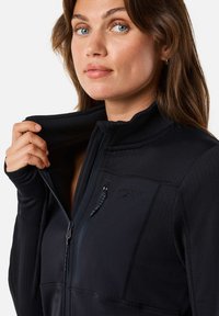 Mustat vetoketjullinen takki korkealla kauluksella, teksturoitua kangasta, sivuvetoketjutasku ja peukalonreiät hihansuissa. Rinnassa logo.