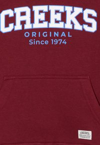 Sweatshirt bordeaux avec texte bleu. Présente "CREEKS ORIGINAL" et "Depuis 1974" imprimés sur le devant. Inclut une poche avant et une étiquette en tissu à l'ourlet.