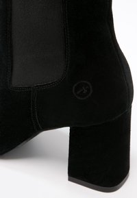 Bottine noire en suède avec un talon carré, comportant un panneau en textile sur le côté et un petit logo à l'arrière. Texture lisse dans l'ensemble.