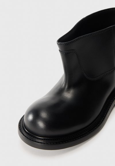 Bota de tobillo de cuero negro con punta redondeada, textura suave y sutil detalle de costura. Presenta una suela de goma baja y duradera.