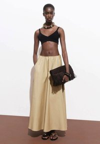 Crni crop top, duga bež suknja s nabranim strukom, smeđa clutch torba s resicama i crne sandale. Ogrlica od velikih perli kao dodatak.