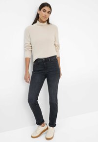 Maglione a collo alto a costine beige abbinato a jeans in denim scuri e sneakers color crema. I jeans presentano una vestibilità classica con cuciture dettagliate.