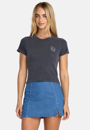 Młoda kobieta z długimi blond włosami, ubrana w dopasowany ciemnoszary krótki t-shirt i niebieską mini spódnicę dżinsową z przednim rozcięciem.