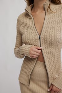 Μπεζ πλεκτό πουλόβερ με κλειστό φερμουάρ, που διακοσμείται με υφασμένο σχέδιο κόμπου και ριμπ μανσέτες. Συνδυάζεται με φούστα σε αντίστοιχο ριμπ σχέδιο.