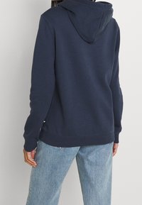 Sweat à capuche bleu marine avec une poche kangourou et une capuche ajustable. Le tissu semble doux avec des poignets et un ourlet côtelés, complété par un jean à coupe ample.