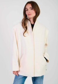 Overmaatse crèmekleurige jas gemaakt van zacht materiaal met een hoge kraag, open voorkant en zijzakken met rits.