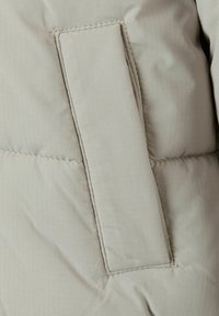 Section d'une doudoune beige clair avec un tissu texturé, des coutures horizontales et un patch rectangulaire en Velcro.