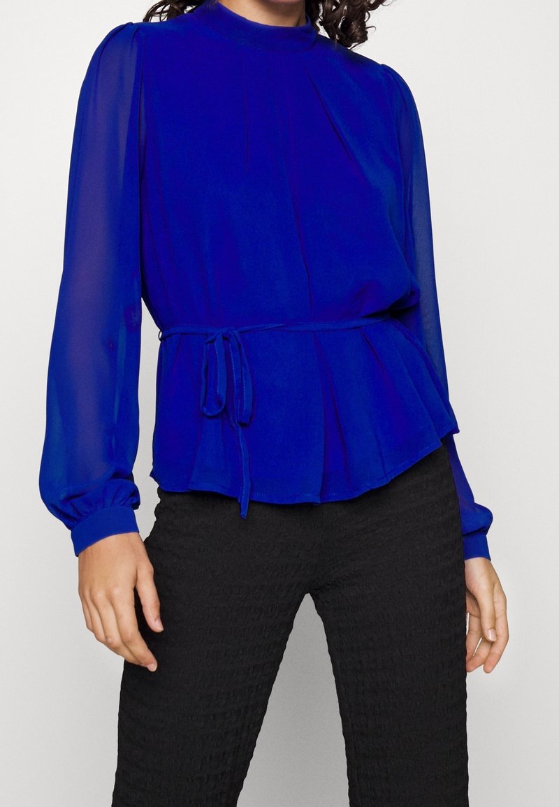 Blusa blu, trasparente, con maniche lunghe, colletto alto e dettagli arricciati in vita. La vita legata accentua la silhouette. Abbinata a pantaloni neri texturizzati.
