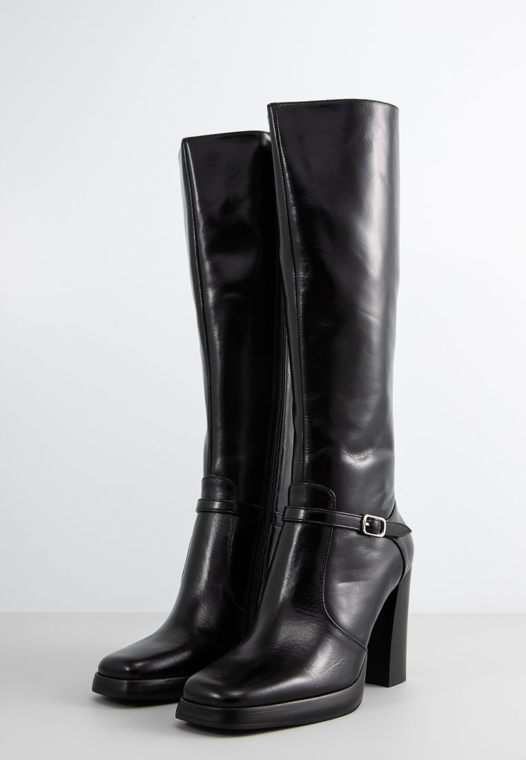 Bottes noires en cuir montantes avec un talon épais, dotées d'une fermeture éclair sur le côté et d'une fine sangle avec une boucle métallique en accent. Texture lisse.