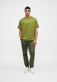 Zöld pamut póló logóval, zöld cargo nadrággal, amely rugalmas mandzsettával rendelkezik, és fehér sportcipővel párosítva. Egyszerű, laza ruházati dizájn.