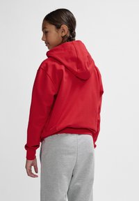 Roter Kapuzenpulli mit lockerer Passform, langen Ärmeln, Kordelzugkapuze und gerippten Bündchen. Kombiniert mit grauen Jogginghosen.