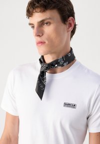 Fekete selyemsál fehér mintákkal a nyak körül, fehér póló fölött viselve, amelyen egy kis fekete "Barbour International" logó található.