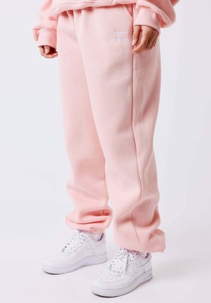 Personne portant un pantalon de survêtement rose poudré avec le logo PXLP et des baskets blanches, debout devant un fond clair uni.