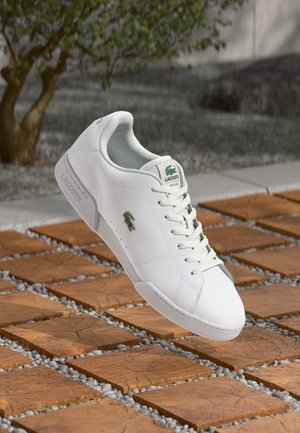 Witte leren sneaker met vetersluiting en groen krokodillologo. Heeft een gladde textuur en een minimalistisch ontwerp op een betegelde achtergrond.