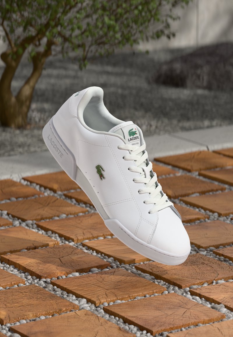 Sapatilhas brancas em pele com atacadores à frente e logótipo de croco verde. Apresentam uma textura suave e um design minimalista sobre um fundo de azulejos.