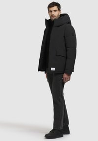 Chaqueta acolchada negra con un cuello alto, cremallera frontal, bolsillos grandes y detalle de logo de goma. Llevada con jeans oscuros y botas negras.