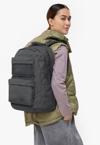 Šedý látkový batoh s několika zipovými kapsami a nastavitelnými popruhy, nošený přes zelenou péřovou vestu a světle fialovou dlouhouchou košili.