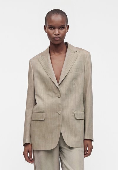 Personne aux cheveux courts portant une veste de costume beige oversize et un pantalon assorti sur un fond blanc uni.