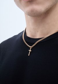 Arany lánc nyaklánc, amely egy kis kereszt medállal díszített, egy sima textúrájú fekete rövidujjú pólón pihen.