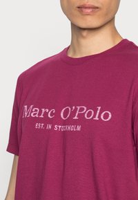 Bordeauxfarvet bomulds T-shirt med korte ærmer, rund hals og hvidt trykt tekst: "Marc O'Polo EST. IN STOCKHOLM" centreret på brystet.