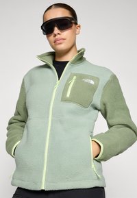 Roheline tooni fleece-mantel, millel on tekstuuriga viimistlus, varustatud lukuga rinna taskuga ja kontrastsete lukkuaktsentidega.