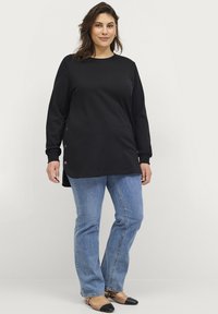 Top negro de manga larga con botones laterales, dobladillo redondeado, combinado con jeans de color azul claro y zapatos planos bicolores, que presenta un ajuste relajado.