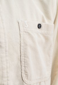 Beige corduroy shirt met een borstzak met zwarte knoop en witte stikselaccenten. De stof heeft een duidelijke ribbelstructuur.
