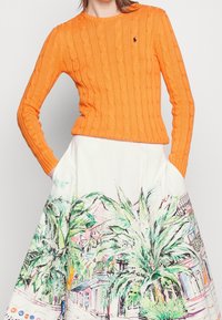 Henkilö, joka on pukeutunut kirkkaan oranssiin neulepuseroon ja valkoiseen hameeseen, jossa on värikkäitä palmun ja rakennuksen kuvituksia, kädet taskuissa.