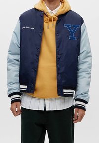 Person som bär en marinblå varsity-jacka med ljusblå ärmar, logotyper för "Yale University" och "Y", senapsgul hoodie, randig vit skjorta och mörka byxor.