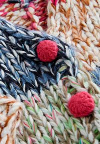 Šarena pletena tkanina s debelom pređom u nijansama plave, ružičaste i bež, ukrašena teksturiranim, okruglim crvenim pomponima.