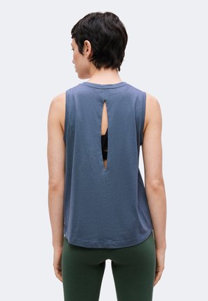 Personne aux cheveux courts et foncés portant un haut bleu sans manches avec une fente verticale au dos et un legging vert foncé, de dos sur un fond uni.