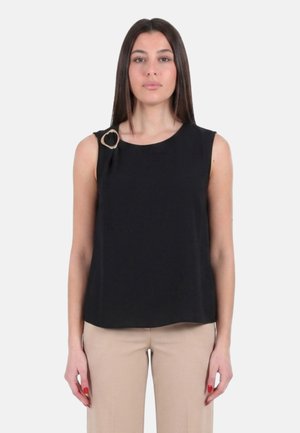 Donna con top nero senza maniche con un anello dorato decorativo sulla spalla sinistra, abbinato a pantaloni beige, in piedi davanti a uno sfondo neutro.