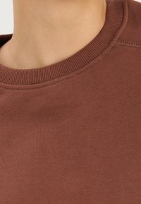 Detailní záběr na krk a rameno osoby oblečené v hnědém svetru s kulatým výstřihem, se viditelnými švy.