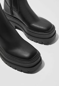 Mustat nahkakumisaappaat, joissa on paksu korokepohja ja kuvioitu pohjakuvio, sileä päällinen pyöreällä kärjellä sekä hienovaraiset ompelukoristeet.